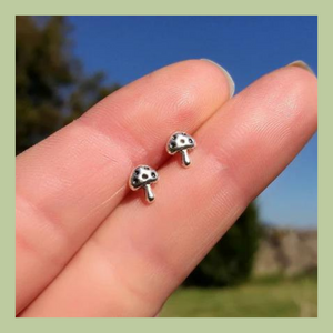 a pair of sterling silver toadstool stud earrings held between fingers in a sunny countryside garden