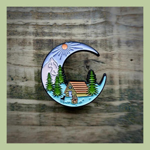 An enamel pin badge with mountains and a campfire with text that says get lost on a bark background