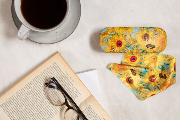 Sunflower-patterned glasses case next to a cup of coffee and an open book on a light surface.