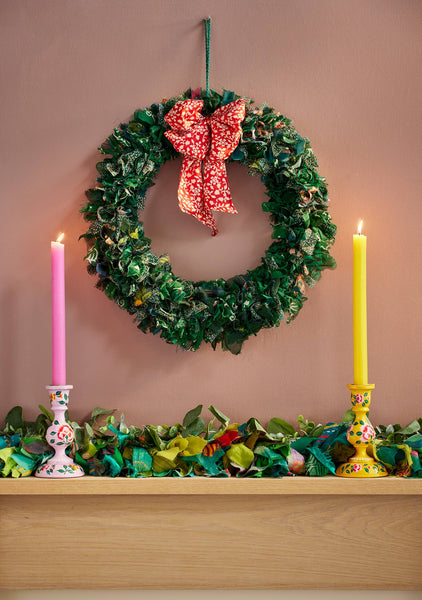 Decorative wreath with a red bow, candles, and garland on a wooden surface against a pink wall.