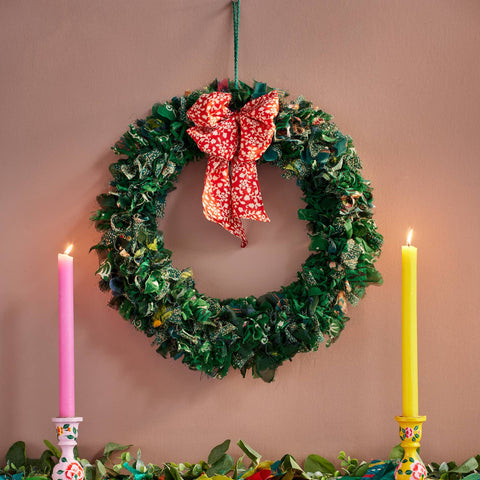 Decorative wreath with a red bow, candles, and garland on a wooden surface against a pink wall.