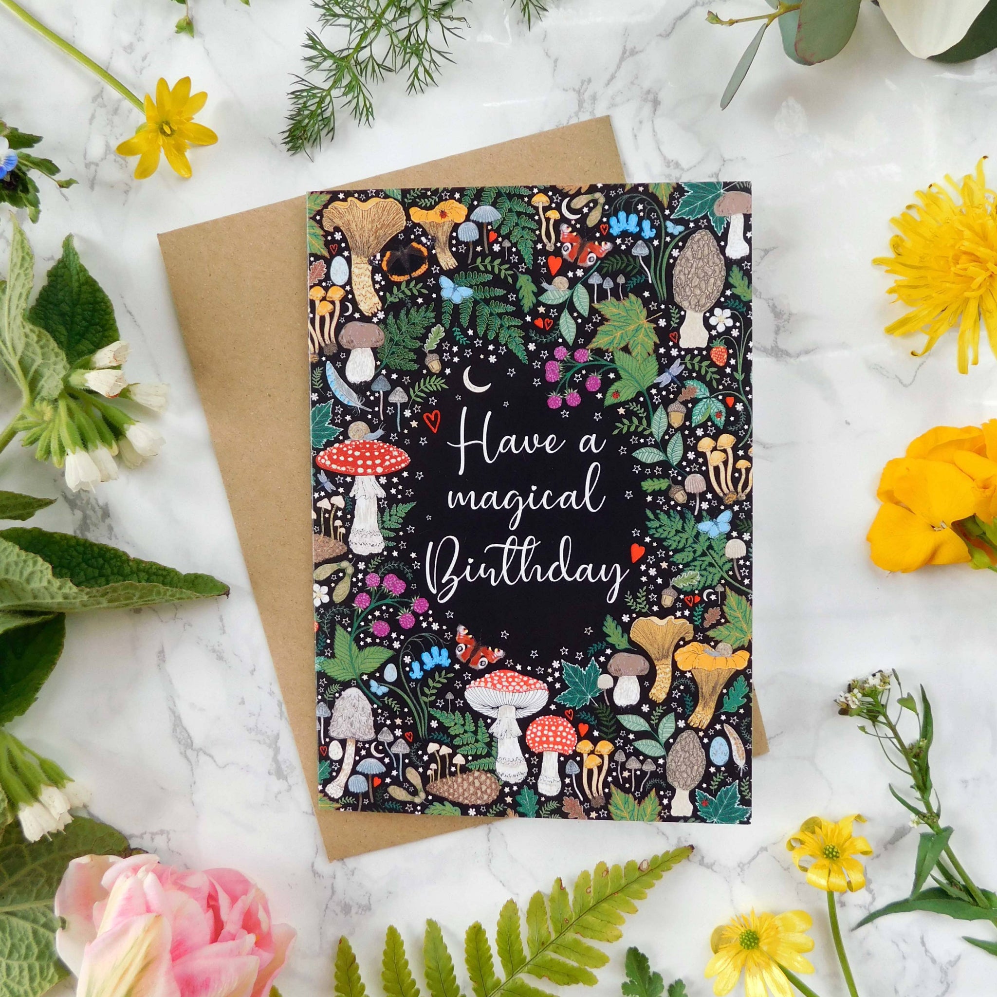 Greeting card with mushroom design and 'Have a magical birthday' text, surrounded by flowers on a marble surface.