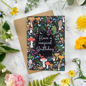 Greeting card with mushroom design and 'Have a magical birthday' text, surrounded by flowers on a marble surface.