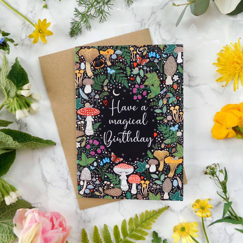 Greeting card with mushroom design and 'Have a magical birthday' text, surrounded by flowers on a marble surface.