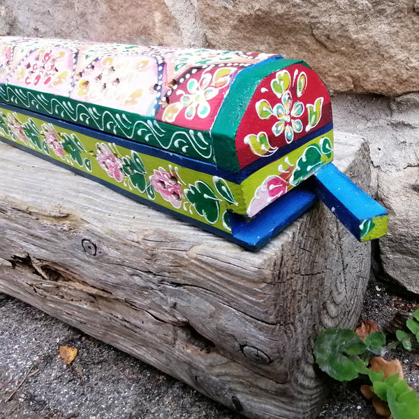 Colorful incense box with floral design on a wooden post against a stone wall.