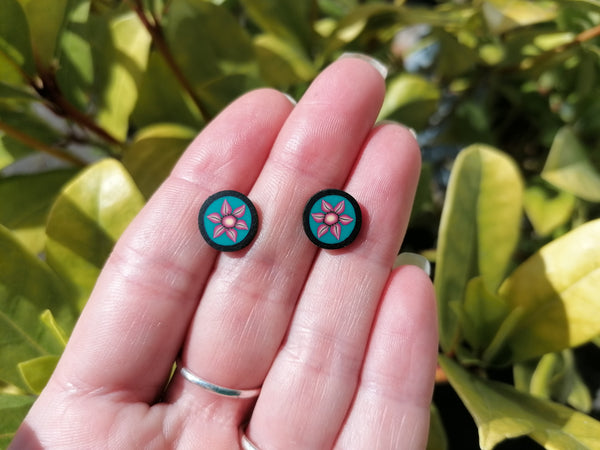 Round earrings with a floral design held between fingers against a leafy green background