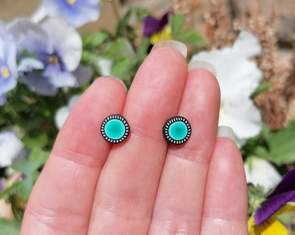 Turquoise stud earrings held between two fingers with a blurred floral background