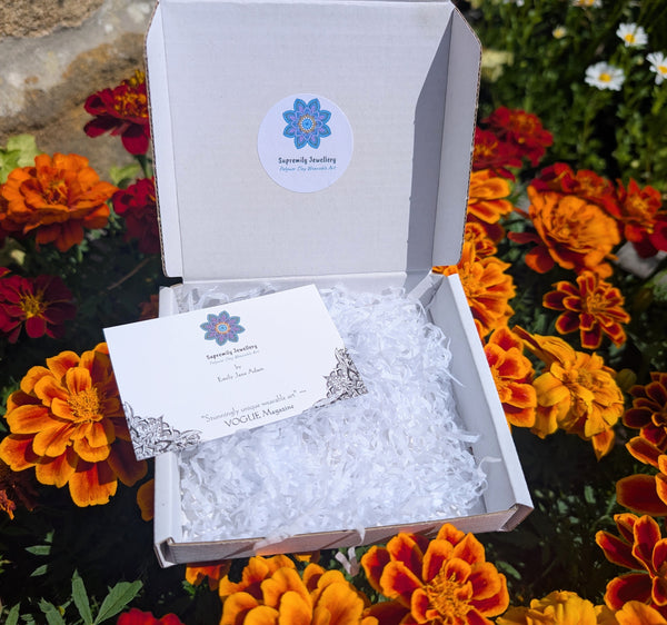 An open white gift box filled with white shredded paper and a branded card, placed outdoors among orange and red marigold flowers. The box lid has a round blue and white logo sticker reading 'Supremily Jewellery'.