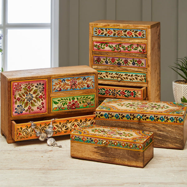 A set of hand-painted wooden jewelry boxes and drawers with intricate floral and geometric patterns in vibrant colors, arranged on a light wooden floor in front of a paneled wall.