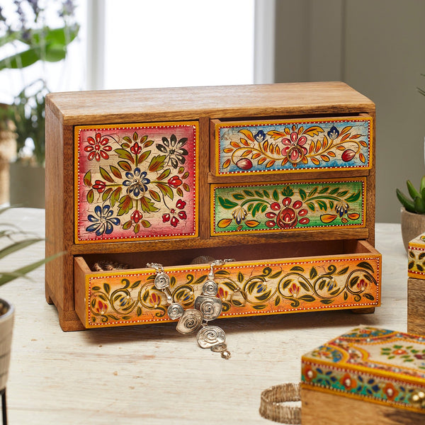 A small hand-painted wooden jewelry box with three drawers, featuring colorful floral and geometric patterns in orange, green, red, and blue. The top drawer is open, revealing silver-toned hanging earrings, and the box is styled on a light-colored tabletop with a blurred window and plants in the background.