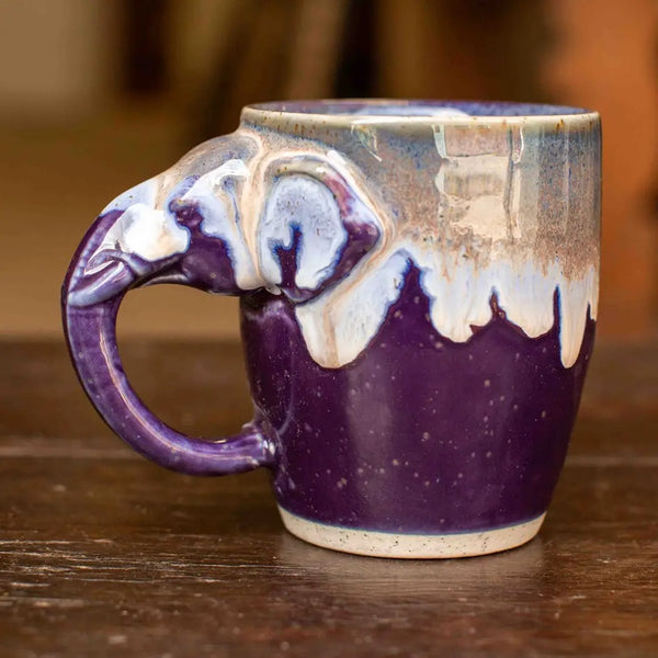 A handmade ceramic mug with a sculpted elephant head handle, featuring a deep purple and white glaze with a dripping effect near the rim, set on a rustic wooden table.