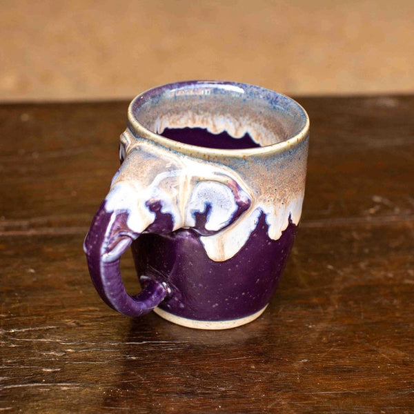A handmade ceramic mug with a sculpted elephant handle, featuring a deep purple glaze with white and beige dripping lava-style patterns, set on a dark wooden table.