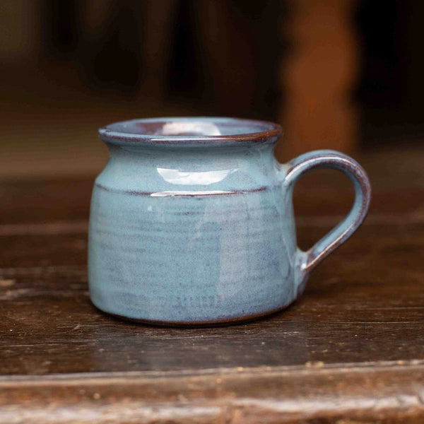 A small handmade ceramic mug with a light blue-green glaze and a dark brown rim, sitting on a rustic wooden surface.