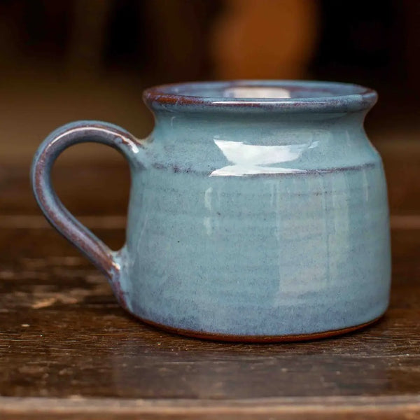 A small ceramic mug with a rounded body, a curved handle, and a rustic glazed finish in light blue with brown clay showing around the rim and base, set on a wooden surface.