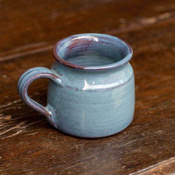 A small handmade ceramic mug with a rounded body, a loop handle, and a wide rim, glazed in a dusty light blue color with a rustic dark brown unglazed edge and rim, sitting on a dark wooden table.