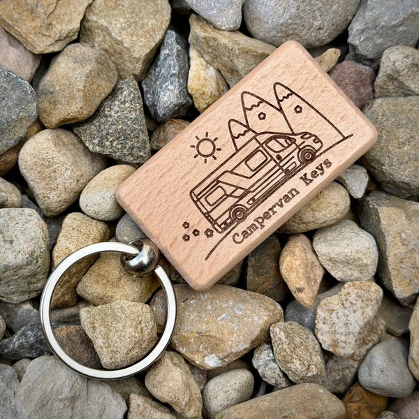 A rectangular wooden keychain with an engraved camper van and trees, labeled 'Campervan Keys,' attached to a silver metal key ring and placed on a bed of gray and beige river stones.