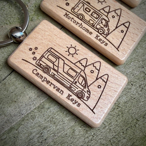 Two engraved wooden camper van keychains on a weathered gray-green wooden surface. The front keychain is engraved with 'Campervan Keys' and a camper van illustration, with a metal key ring partially visible.