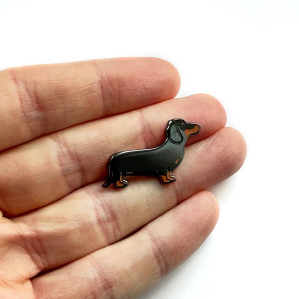 Black dachshund-shaped pin held between fingers on a white background