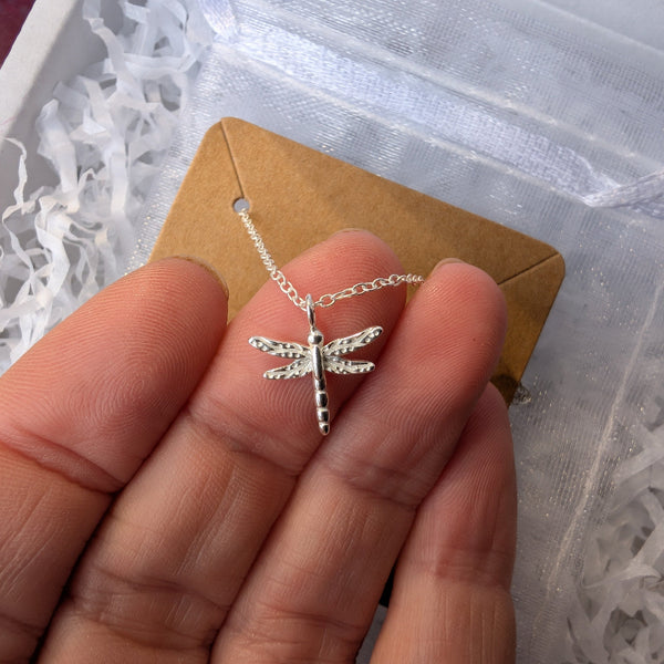 Silver dragonfly necklace on a cardboard card held by a hand, with a white textured background.
