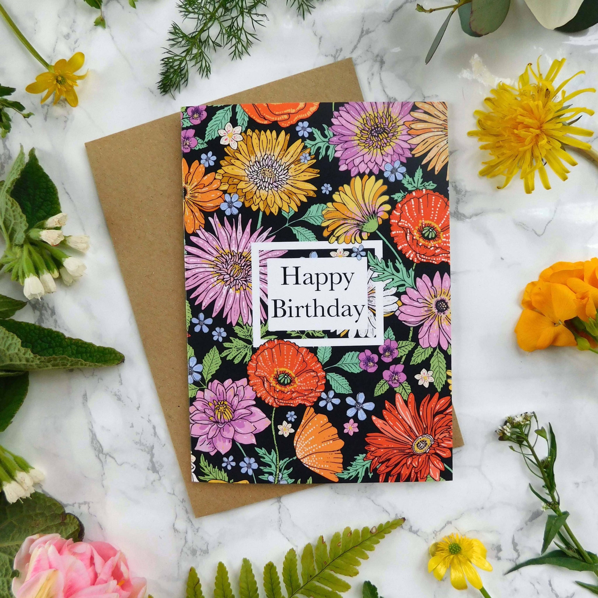 Birthday card on a marble surface with floral arrangement. The card says happy birthday and has colourful illustrated flowers on the front.