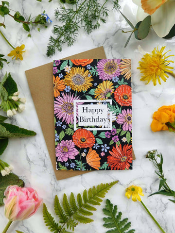 Birthday card on a marble surface with floral arrangement. The card says happy birthday and has colourful illustrated flowers on the front.