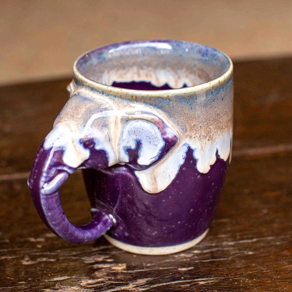 A handmade ceramic mug with a sculpted elephant head handle, featuring a glossy purple and white glaze with a natural brown-rust gradient near the rim, resting on a rustic wooden table.