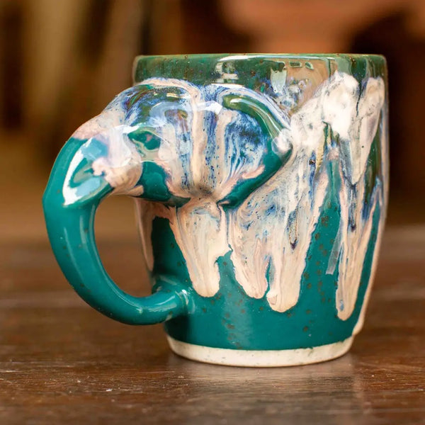 A handmade ceramic mug with a sculpted elephant head handle, featuring a glossy teal green glaze with dripping white and blue paint details, set on a wooden surface.