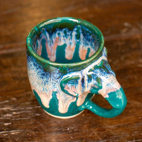 A ceramic mug shaped like an elephant, featuring a glossy teal and blue glaze with white dripping accents, set on a rustic dark wood surface.