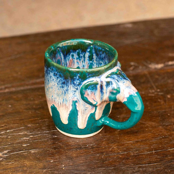 A ceramic elephant-shaped mug with a glossy teal, blue, and white drip glaze, resting on a rustic dark wood surface.