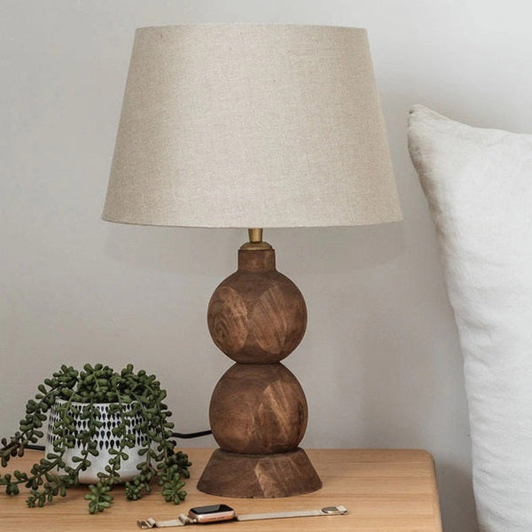 Wooden lamp with beige shade on a wooden nightstand next to a pillow and plant.