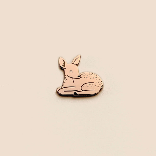 A small enamel pin shaped like a resting deer or fawn, shown on a plain warm beige background.