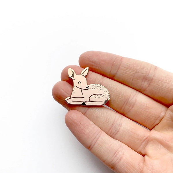 A small deer-shaped enamel pin held between a person's fingers against a plain white background.