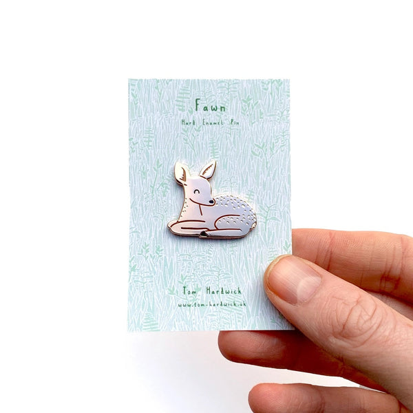 A hand holding a light blue card displaying a small enamel pin shaped like a sleeping deer. The card has green text that reads 'Fawn' and 'rest content in peace', and the pin itself is pale pink with a thin metallic outline.
