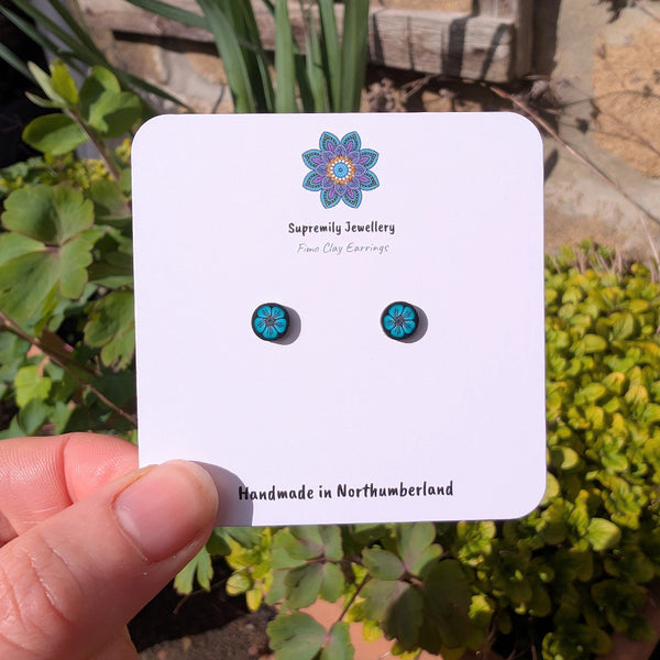 Pair of green floral earrings on a Supremily Jewellery card held in front of green plants.