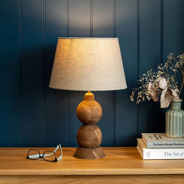 Table lamp with a wooden base and beige shade on a wooden surface against a dark blue paneled wall.