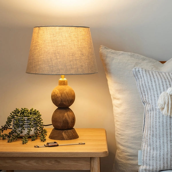 Wooden lamp on a bedside table with a plant and remote control, next to a bed with pillows.