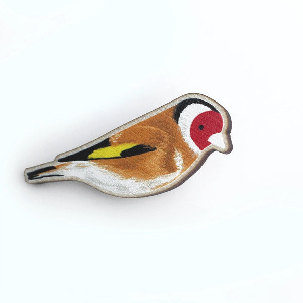 A bird-shaped enamel pin depicting a goldfinch, featuring brown, white, red, yellow, and black details on a silver-toned metal outline, set against a plain white background.
