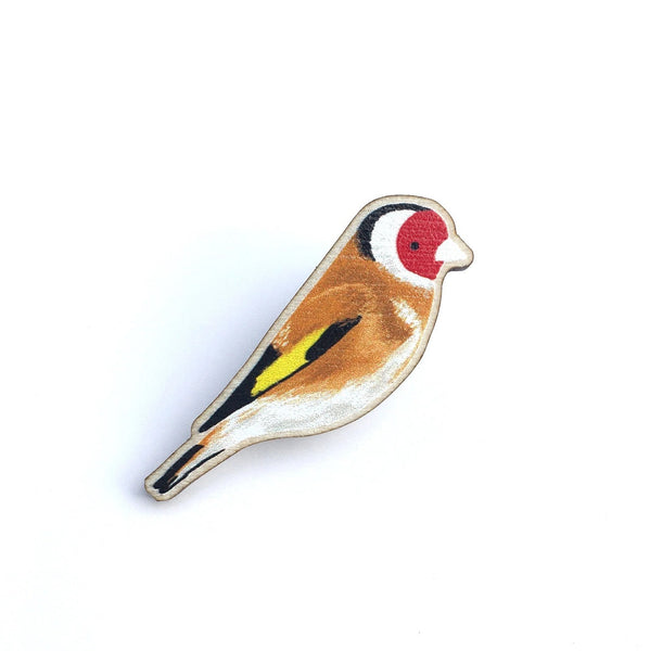 A small bird-shaped enamel pin on a plain white background. The pin depicts a goldfinch with a brown body, white underbelly, red face, black and white eye stripe, yellow wing accent, and black tail details.