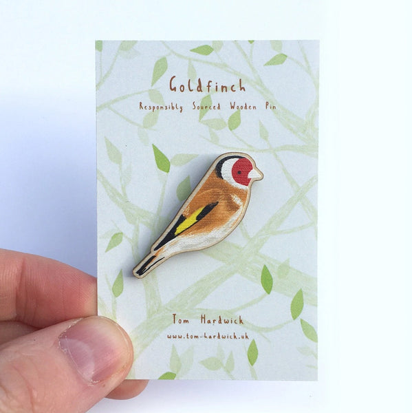 A goldfinch bird enamel pin by Tom Hartwick, displayed on its branded backing card and held by a person's hand against a white background.