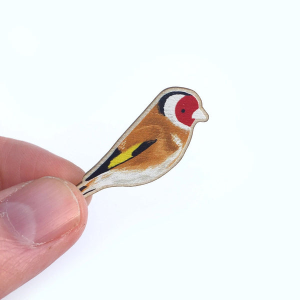 A small bird-shaped enamel pin or charm, painted like a goldfinch with a red face, black and white stripe, yellow wing, and brown body, held between two fingers against a plain white background.