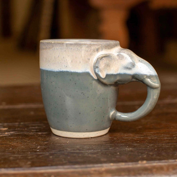 A handmade ceramic mug with a sculpted elephant head handle, featuring a speckled slate blue-gray glaze on the lower body and a matte off-white glazed top and handle, resting on a rustic wooden table.