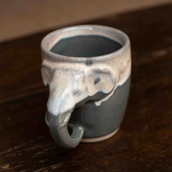 A ceramic mug shaped like an elephant head, featuring a dark gray body with a glossy white and beige dripping glaze, resting on a dark wooden surface.
