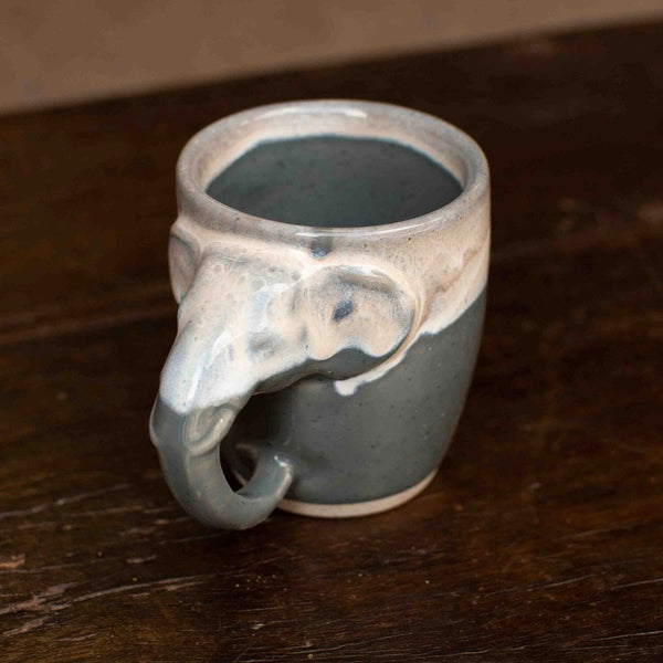 A handmade ceramic mug shaped like an elephant head, featuring a matte slate gray body and a glossy white glazed rim, resting on a dark wooden surface.