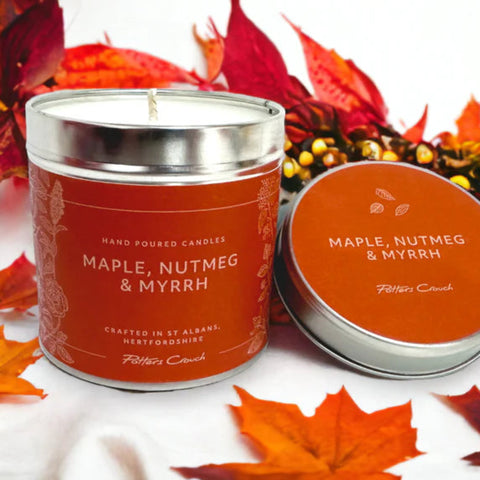A candle with a maple nutmeg and myrrh scent, in a brown tin with a lid, displayed on a white surface with red leaves around it. The label on the candle indicates the scents and the brand Potters Crouch.
