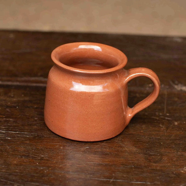 A small terracotta-colored ceramic mug with a wide rim and a curved handle, resting on a dark wooden tabletop.