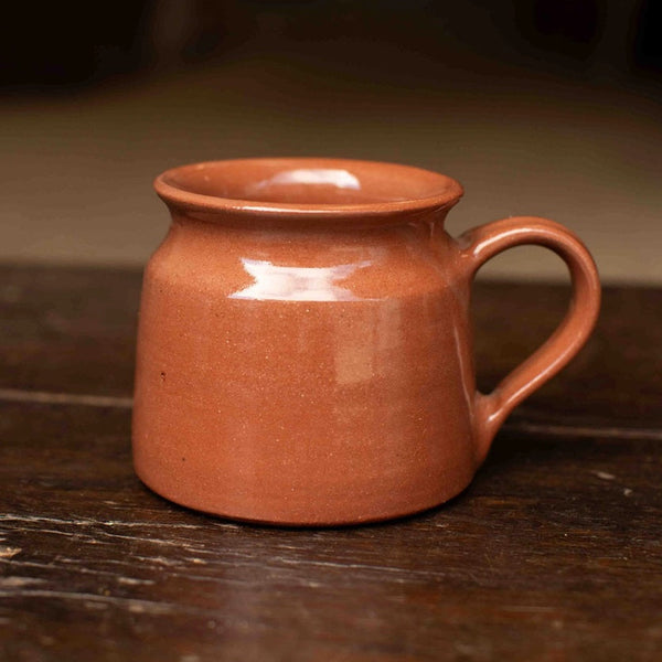 A small terracotta-colored ceramic mug with a wide rim and a curved handle, resting on a dark wooden surface with a softly blurred background.