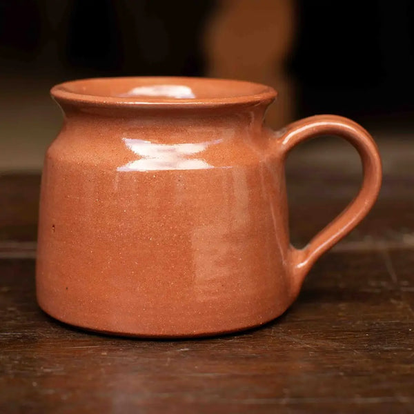 A small handmade ceramic mug with a rounded body and a curved handle, glazed in a warm terracotta orange-brown color, resting on a dark wooden surface.