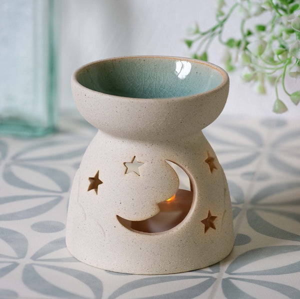 A ceramic wax warmer with a speckled cream exterior, featuring a crescent moon and star cutout design, and a glazed light blue interior. A small tea light candle is placed inside the warmer, which sits on a patterned surface with a softly blurred background.
