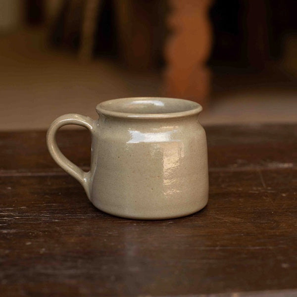A small handmade ceramic mug with a handle, featuring a matte light beige glaze, resting on a dark rustic wooden table.