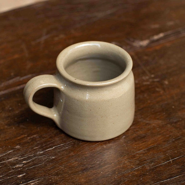 A small ceramic mug with a rounded body and a loop handle, finished in a speckled light gray glaze, resting on a dark wooden table.